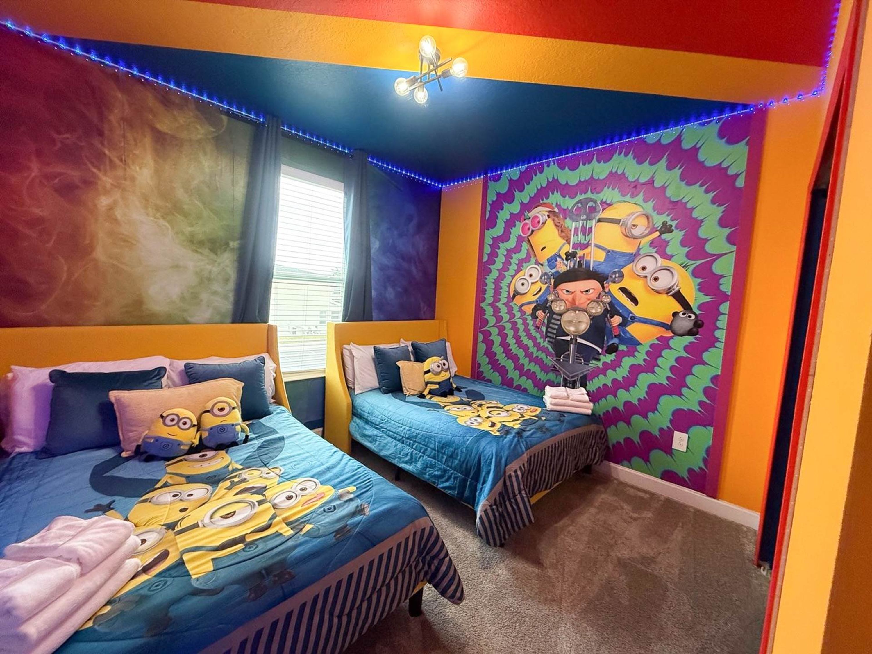 A bedroom with two beds, each with a Minion-themed duvet. The room features a colorful mural on the wall, a window with blinds, and blue LED strip lights.
