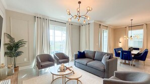 A modern living room with a grey sofa, two armchairs, a coffee table, and a dining area with blue chairs and a chandelier. - Upstay Luxury Living w Pool Hot Tub Cinema (Davenport)