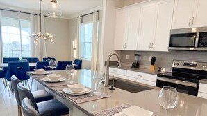 A modern kitchen with white cabinets, a stainless steel microwave, and an oven. There is a dining area with blue chairs and a long table set for four. - Upstay Luxury Living w Pool Hot Tub Cinema (Davenport)