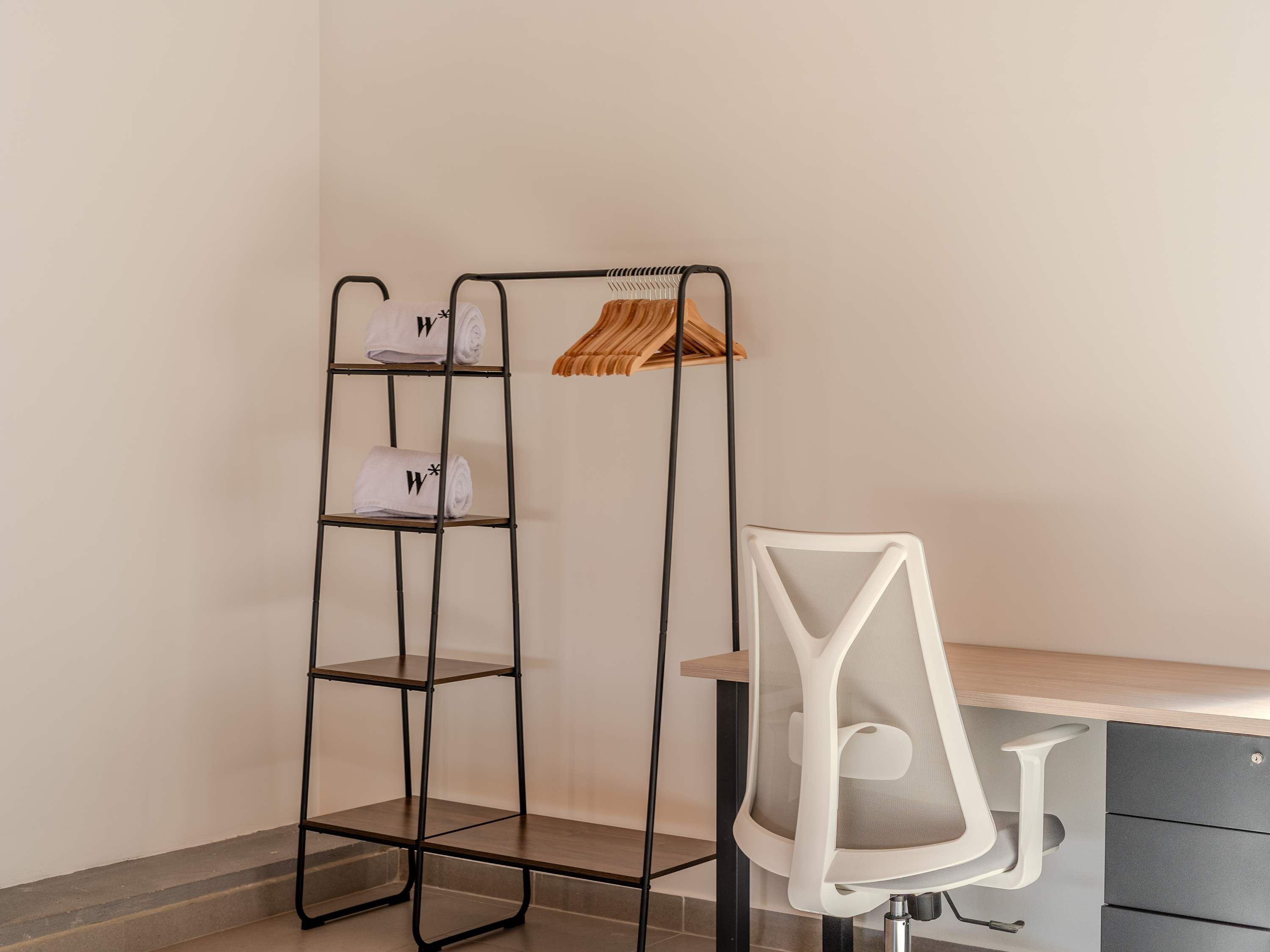 Une étagère métallique avec des serviettes et un cintre en bois, placée à côté d’un bureau muni d’une chaise et de tiroirs.
