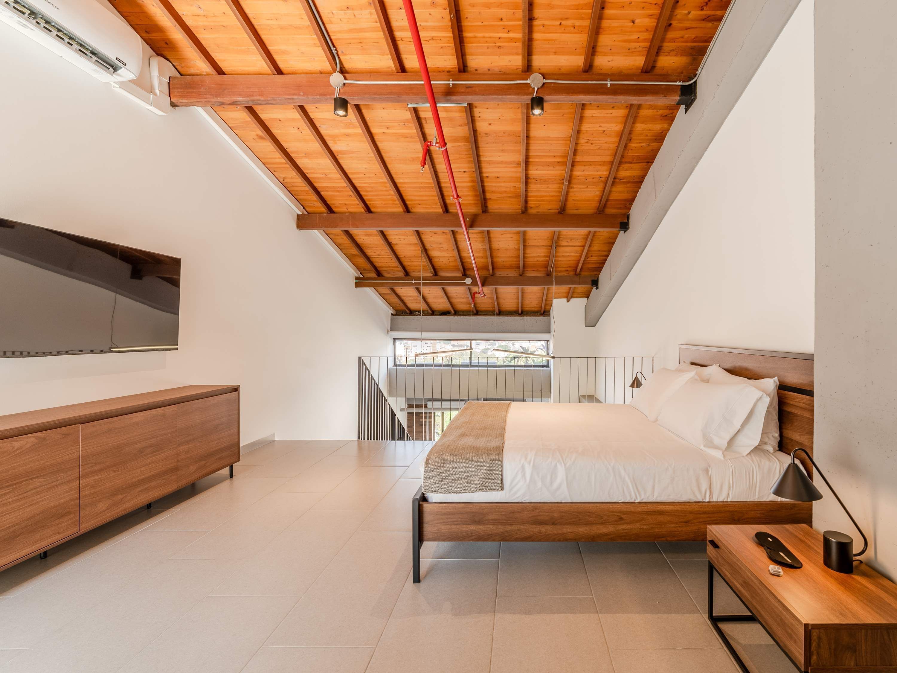 A modern bedroom with a wooden ceiling, a bed, a bedside table, a nightstand, a TV mounted on the wall, and a view of the outdoors.
