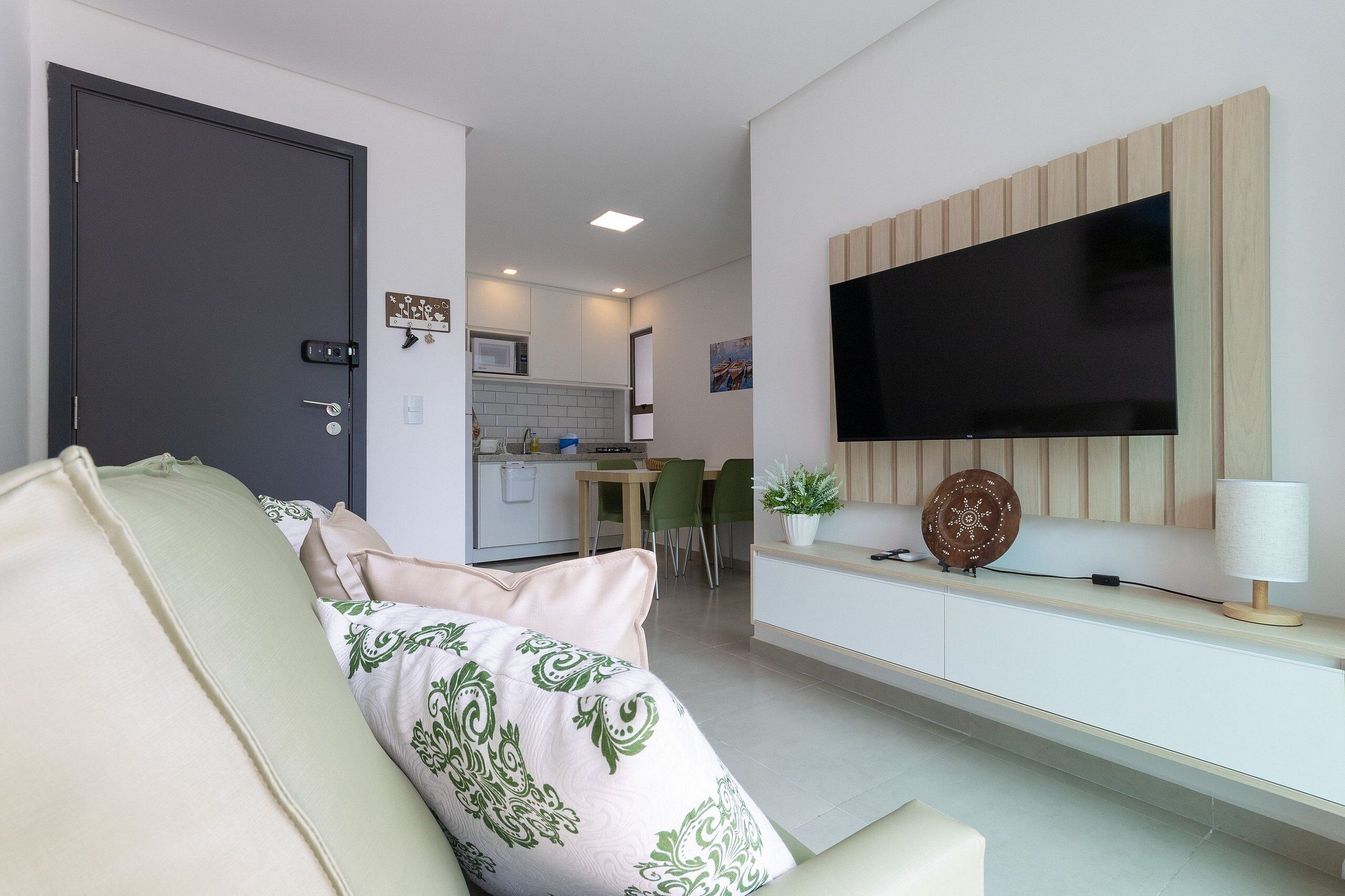 A modern living room with a sofa, a flat-screen TV mounted on a wooden panel, a dining area with a table and chairs, and a kitchen in the background.