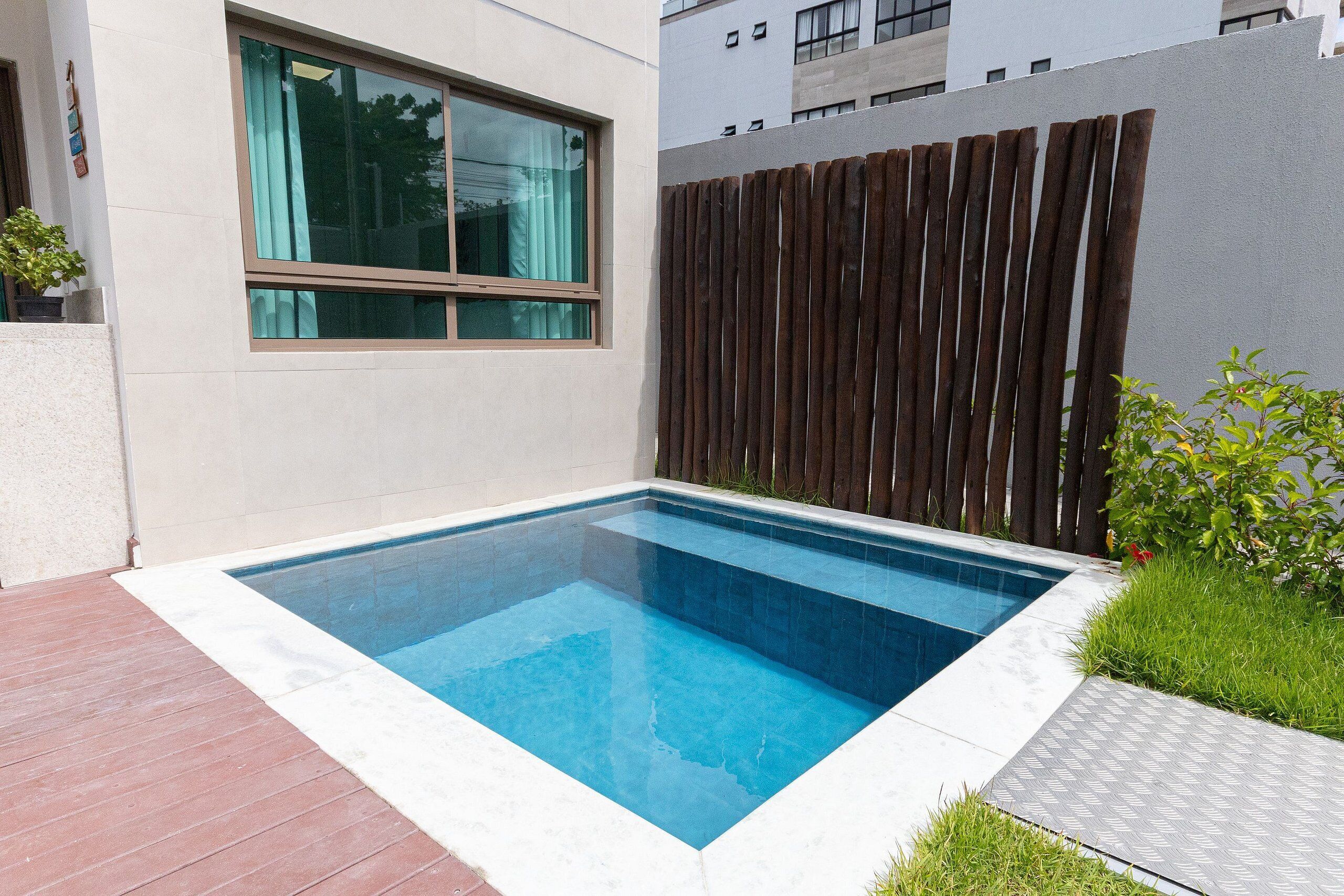 A rectangular swimming pool with a clear blue water surface, surrounded by a white tiled deck and a wooden fence.