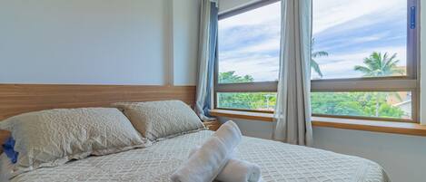 A neatly made bed with a patterned bedspread, a wooden headboard, and a view of palm trees through a window with curtains.