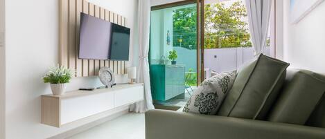 Una sala de estar moderna con un televisor de pantalla plana montado en un panel de madera, un sofá con cojines estampados, una pequeña mesita de noche con una planta y un portón corredizo de vidrio que da a un área exterior.