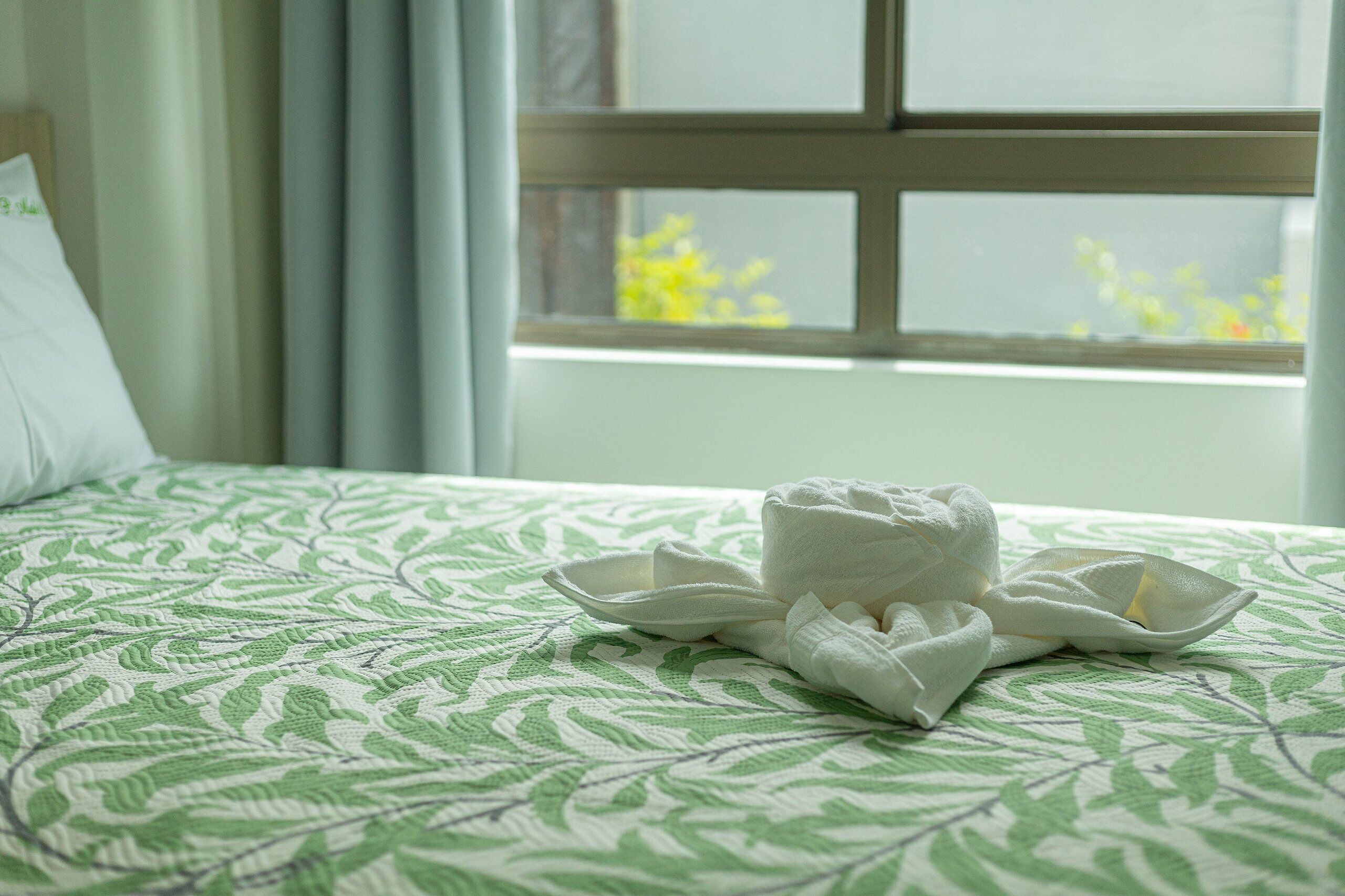 Una cama con un edredón estampado con hojas verdes y una toalla blanca doblada con cuidado.