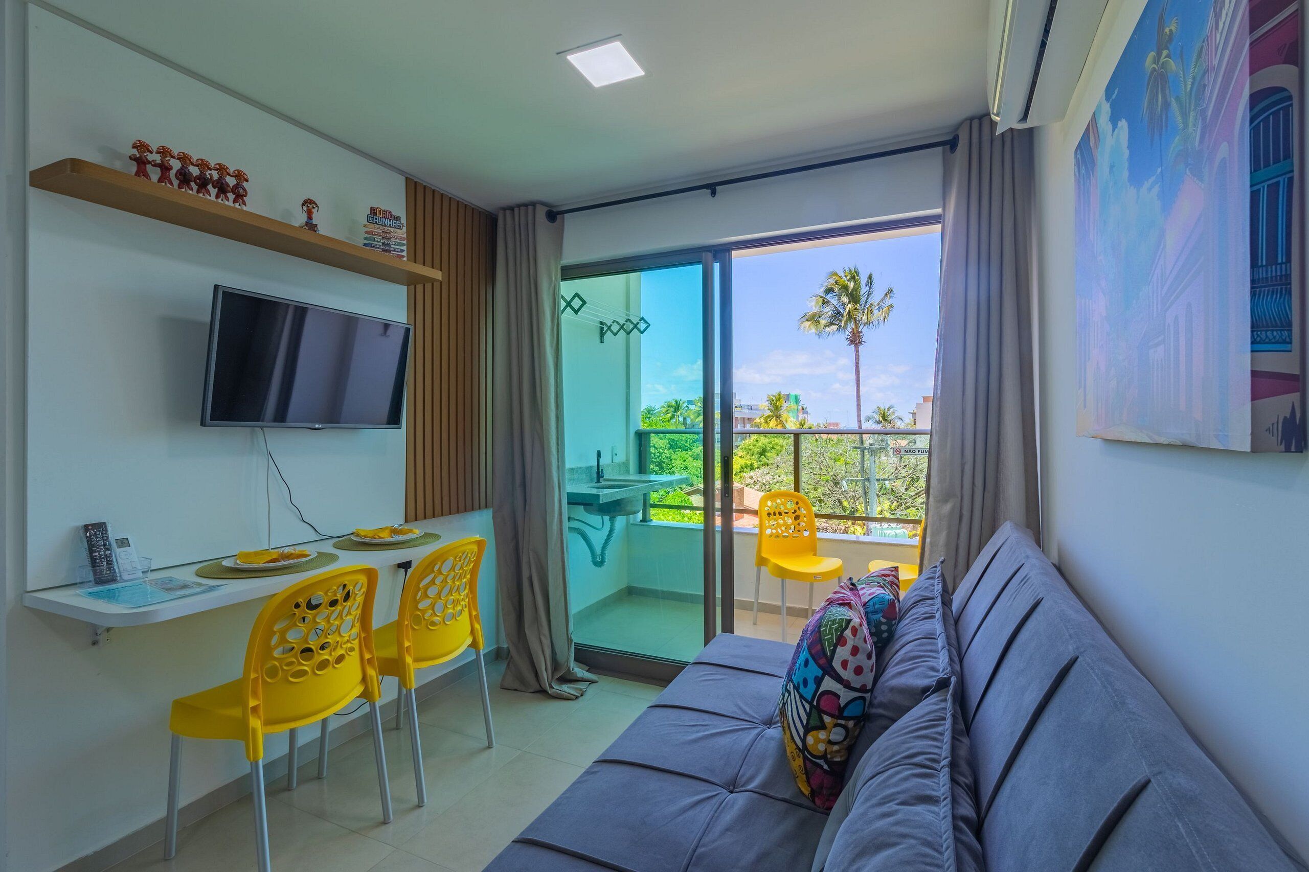 A modern room with a sofa, a TV, a dining table with chairs, a balcony with a sink, and a painting on the wall.