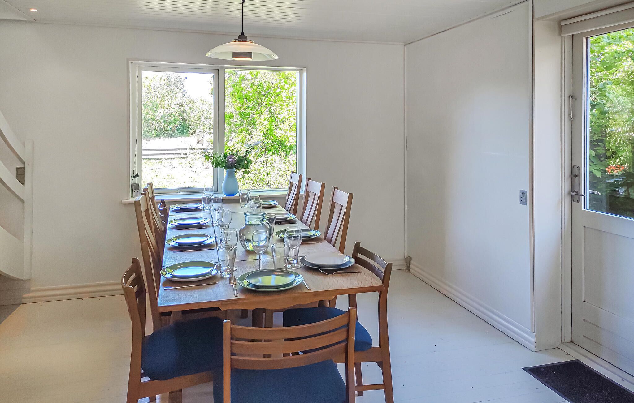 A dining area with a long table set for six, surrounded by wooden chairs with blue cushions. A vase with flowers is on the table, and there is a window providing natural light.