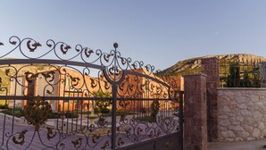 Ein schmiedeeisernes Tor mit aufwendigen Verzierungen, das zu einem Gebäude mit einer Steinmauer und einer Bergkulisse führt.