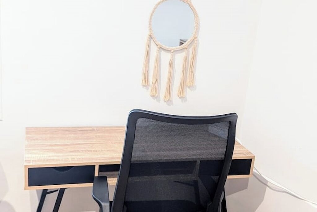 Een werkplek met een houten bureau, een zwarte bureaustoel en een macramé decoratie aan de muur.