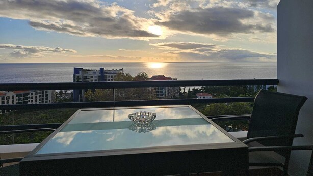 A balcony with a glass table and chair, overlooking a cityscape and the sea during sunset. - Sea View Forum Wi-Fi, AC & Parking 1 minute from Forum Madeira (Funchal)