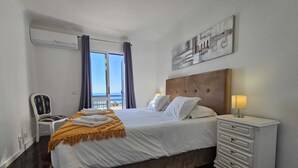 A hotel room with a bed, a nightstand with a lamp, a chair, and a view of the sea through large windows. - Sea View Forum Wi-Fi, AC & Parking 1 minute from Forum Madeira (Funchal)