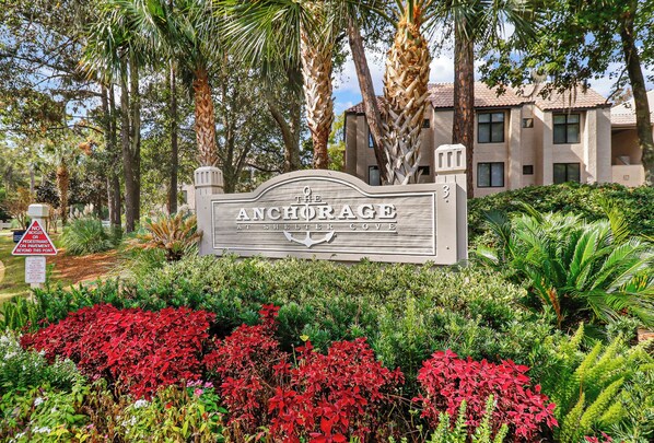 A sign for 'The Anchorage at Shelter Cove' with a no bicycles or pedestrians on pavement sign, surrounded by lush greenery and palm trees.