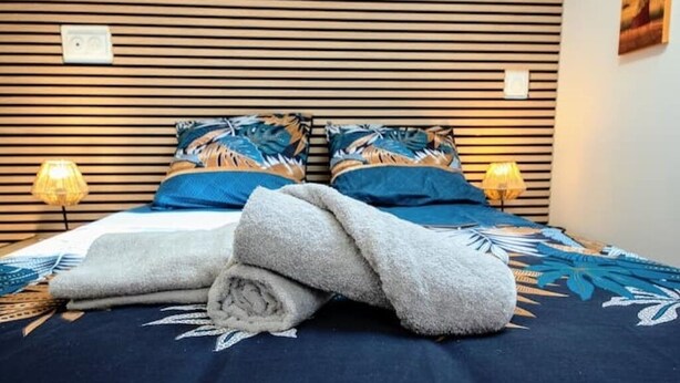 A neatly made bed with a blue and white patterned bedspread, two pillows with tropical designs, and two bedside lamps. - Studio 4 - Bright Cocoon  (Gond-Pontouvre)