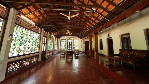 A spacious corridor with a high wooden ceiling, large windows with intricate designs, and traditional wooden furniture.