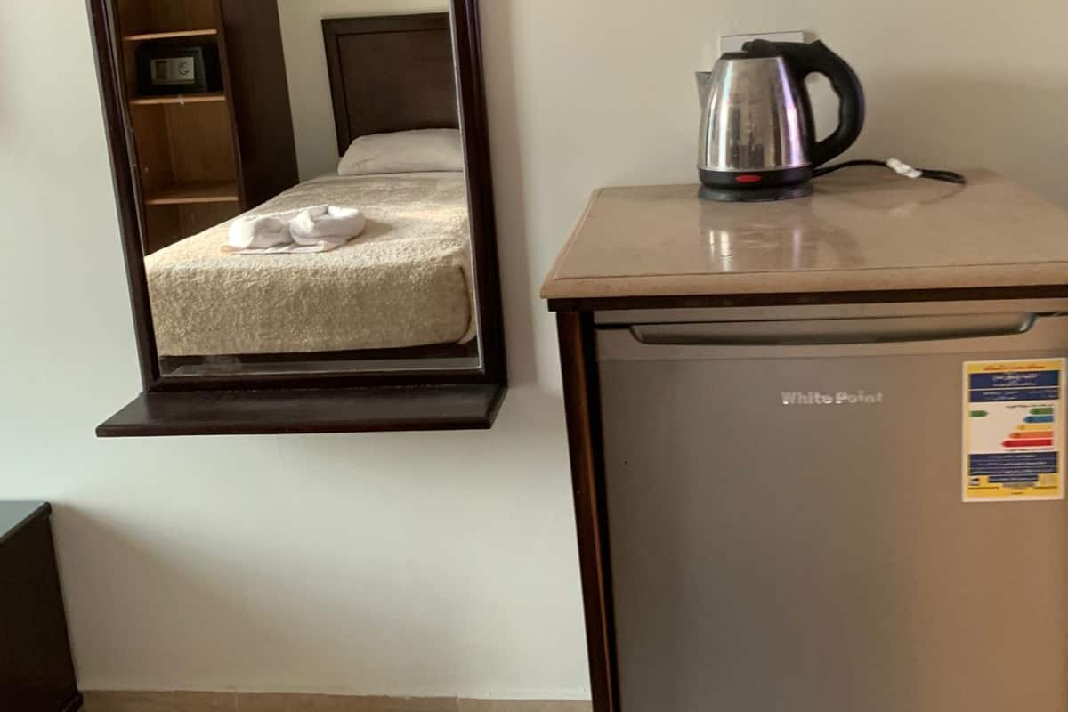 Ein Zimmer mit einem Bett, einem kleinen Tisch mit Wasserkocher, einem Kühlschrank und einem großen Spiegel, der einen Kleiderschrank und eine Tür reflektiert.