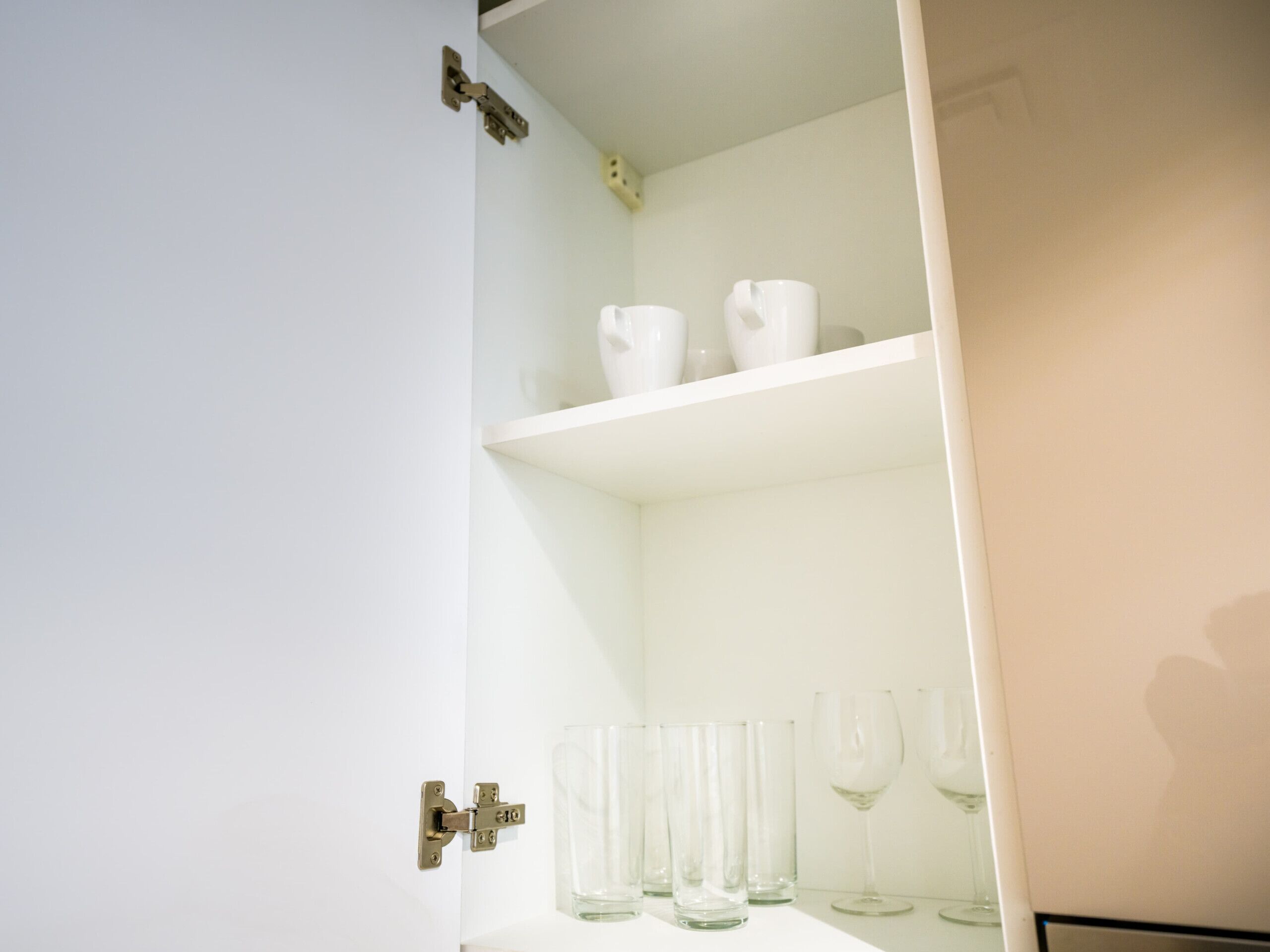 Un armadietto bianco con ripiani che contengono bicchieri trasparenti e oggetti in ceramica bianca.