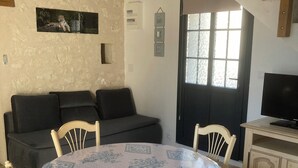 A dining area with a round table covered in a floral tablecloth, surrounded by wooden chairs. There is a black sofa against the wall and a wooden cabinet with a television on top.