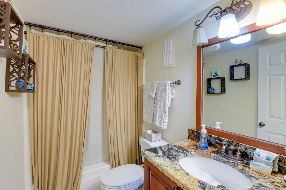 Un baño con inodoro blanco, cortinas color beige, un perchero, un lavabo con encimera de mármol y un espejo con luz.