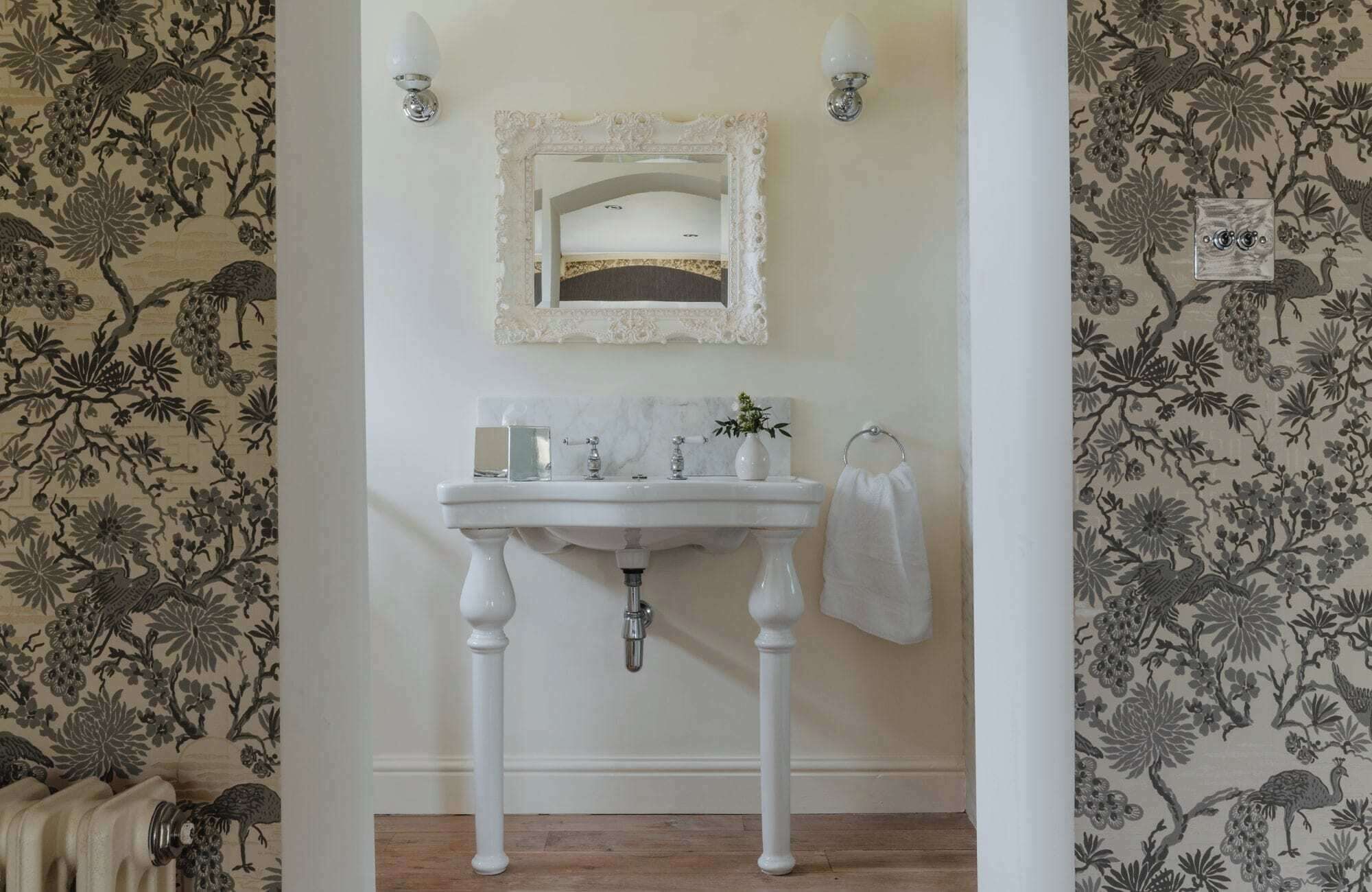 A bathroom with a white pedestal sink, a mirror above it, and a floral wallpaper pattern on the walls.