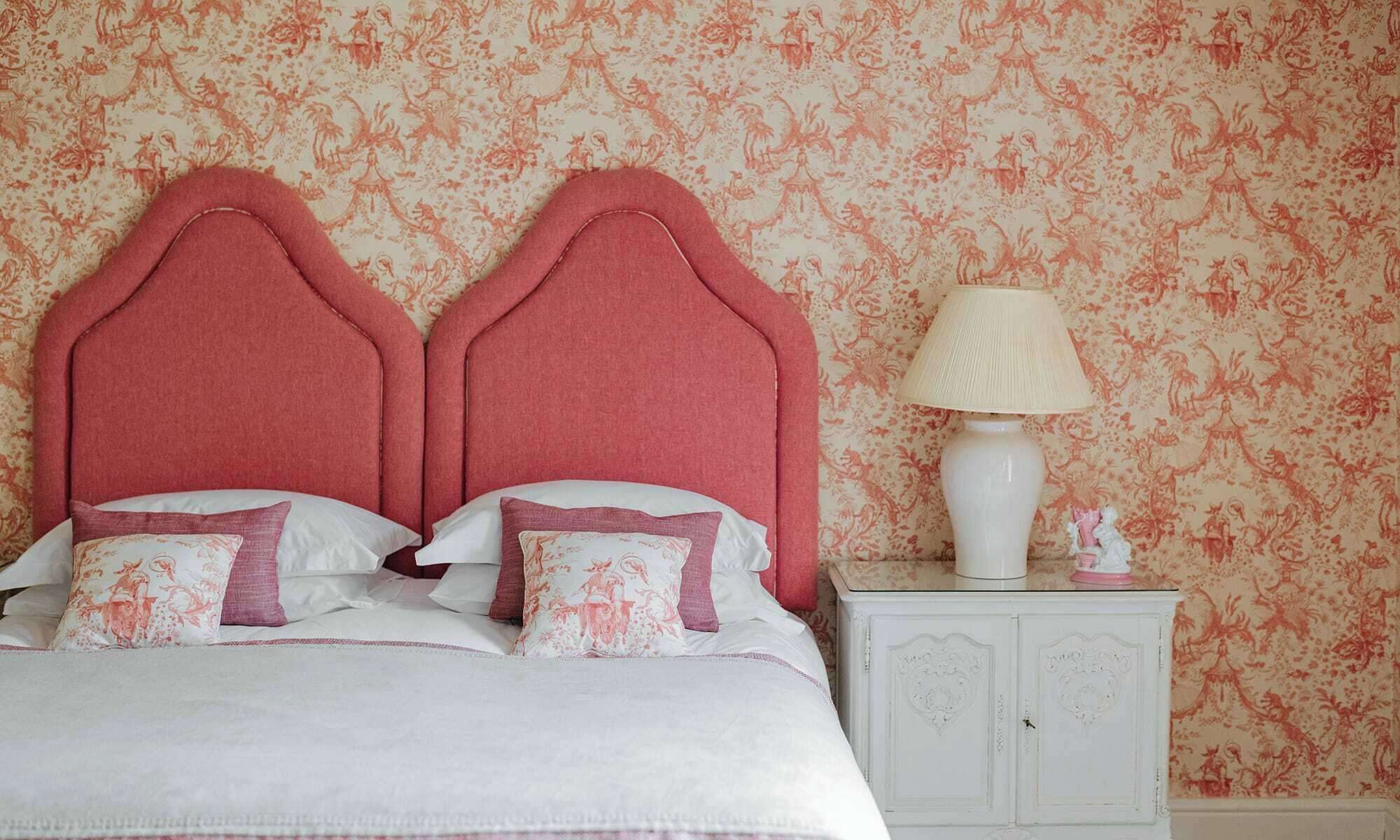 A bed with a pink headboard, white bedding, and decorative pillows. There is a white nightstand with a lamp and a small statue. The wall has a floral patterned wallpaper.