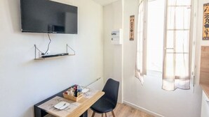 A room with a flat-screen TV mounted on the wall, a wooden table with a chair, and a window with sheer curtains. - Studio 3 - Cocon Douillet : Le Confort  (Gond-Pontouvre)