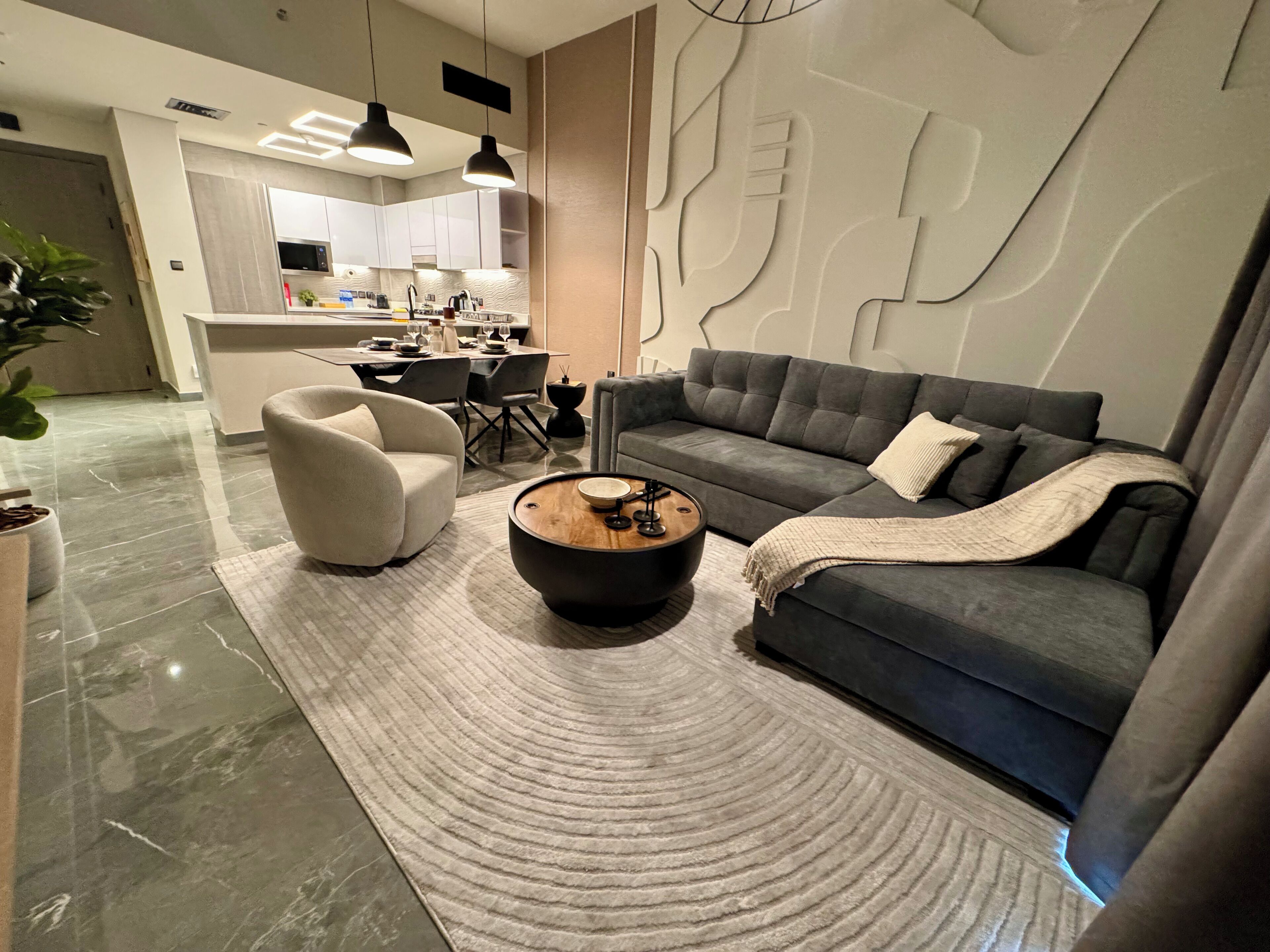 A modern living room with a grey sofa, a white armchair, and a coffee table. There is a kitchen in the background with white cabinets and a dining area.
