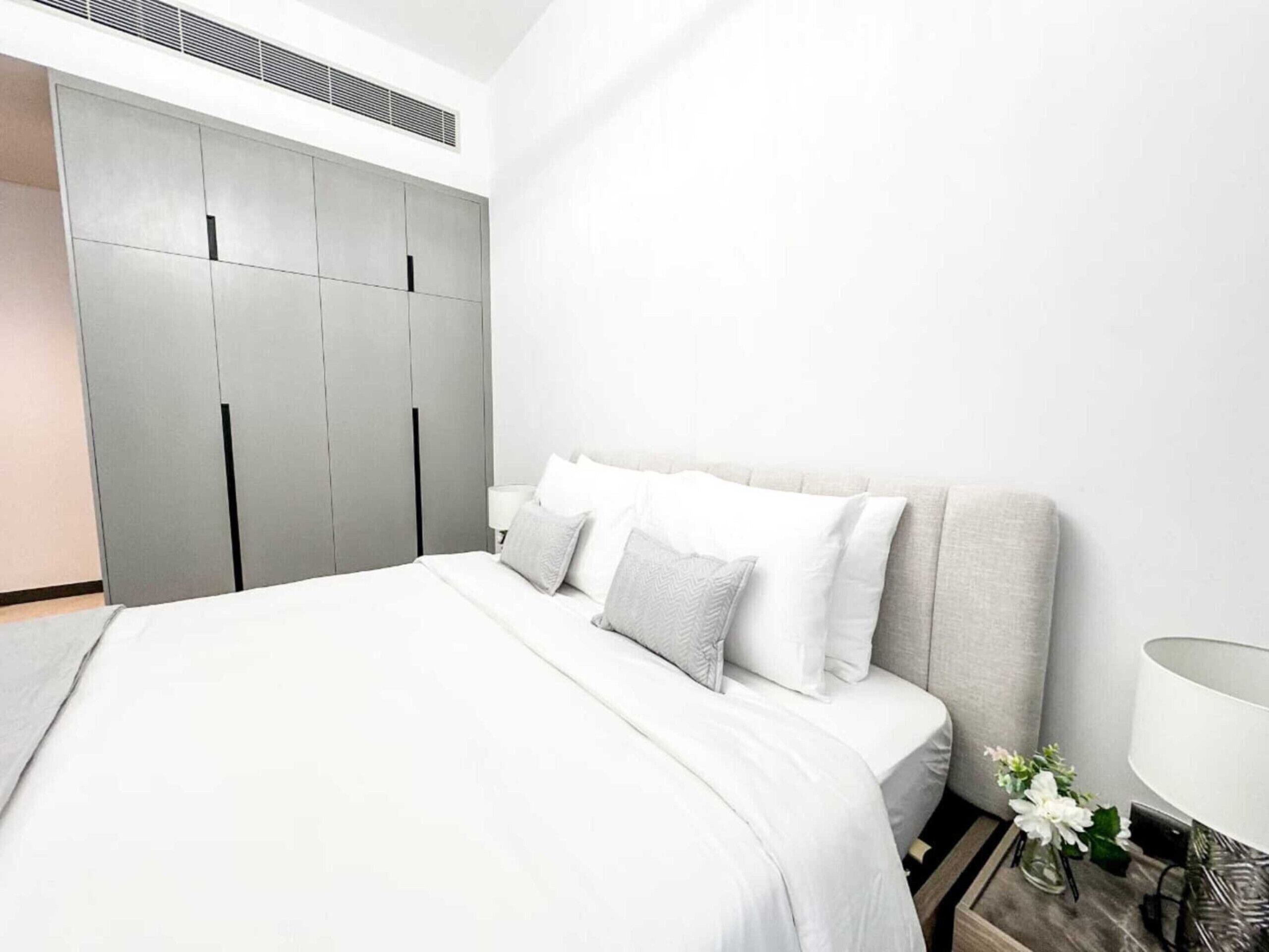 A modern bedroom with a large bed, grey wardrobe, and a bedside table with a lamp and flowers.