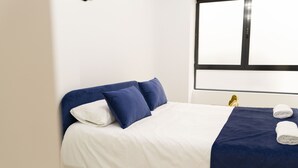 A neatly made bed with white and blue bedding, a blue throw blanket, and two rolled towels on a dark blue bedspread. - Forata Lofts, Room 3 (Valencia)