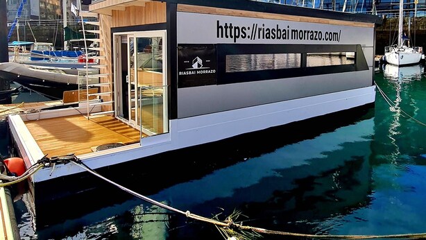 A modern floating structure with a website URL displayed, docked in a marina with buildings and palm trees in the background. - Floating apartment in the nautical Vigo ferry dock Cies Islands and Cangas (Vigo)