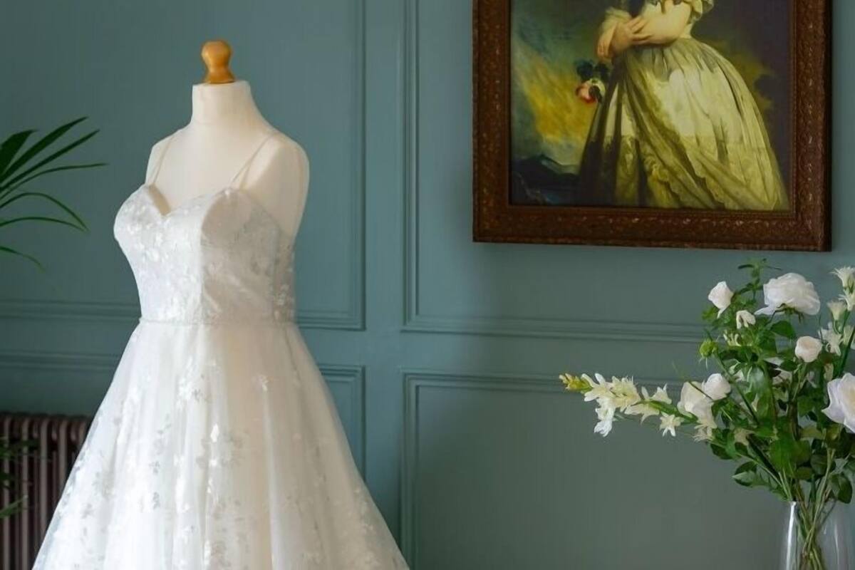 A mannequin in a white wedding dress with lace details, positioned in front of a wall with a framed portrait and a vase of white flowers.