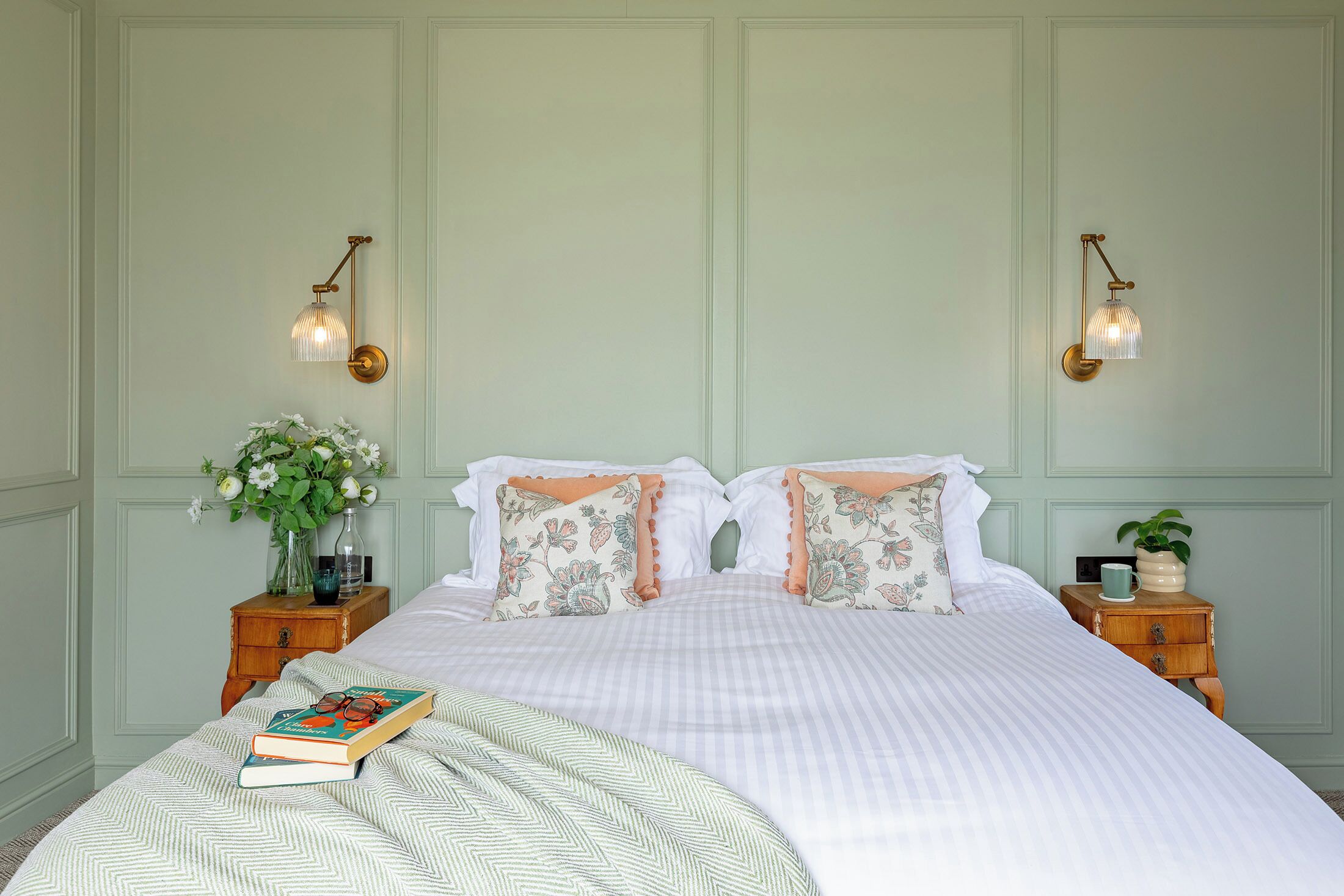 Una cama bien hecha con almohadas florales, un libro y un jarrón de flores sobre una mesita de noche.