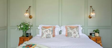 Una cama bien hecha con almohadas florales, un libro y un jarrón de flores sobre una mesita de noche.