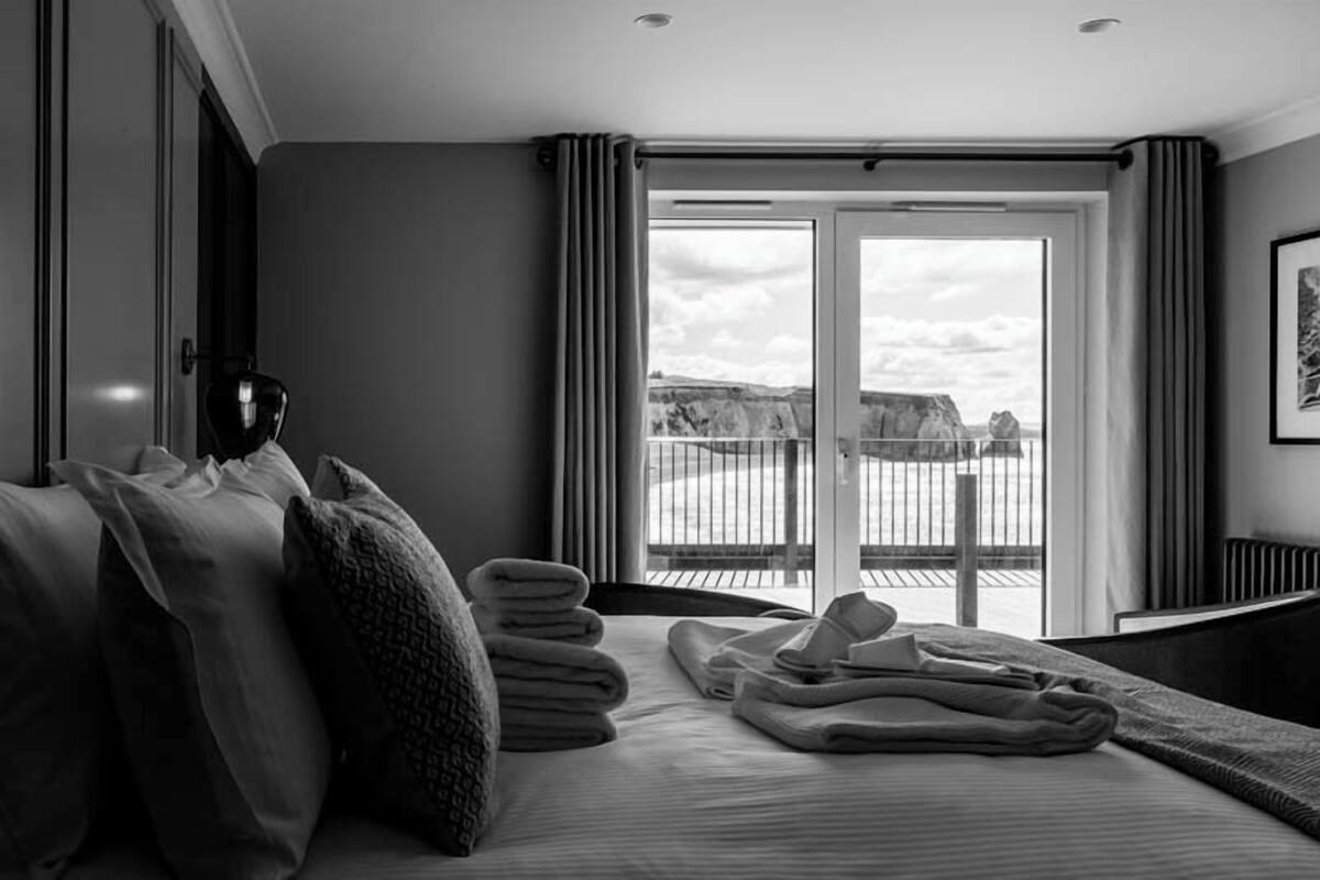 A hotel room with a bed, a side table, a window with curtains, and a view of the outdoors.