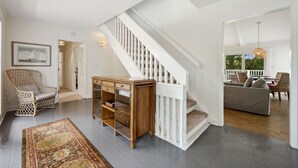A spacious living area with a staircase, a wooden cabinet, a patterned rug, and wicker furniture. - Hellenic Haven 6 Bedroom Home (Georgetown)