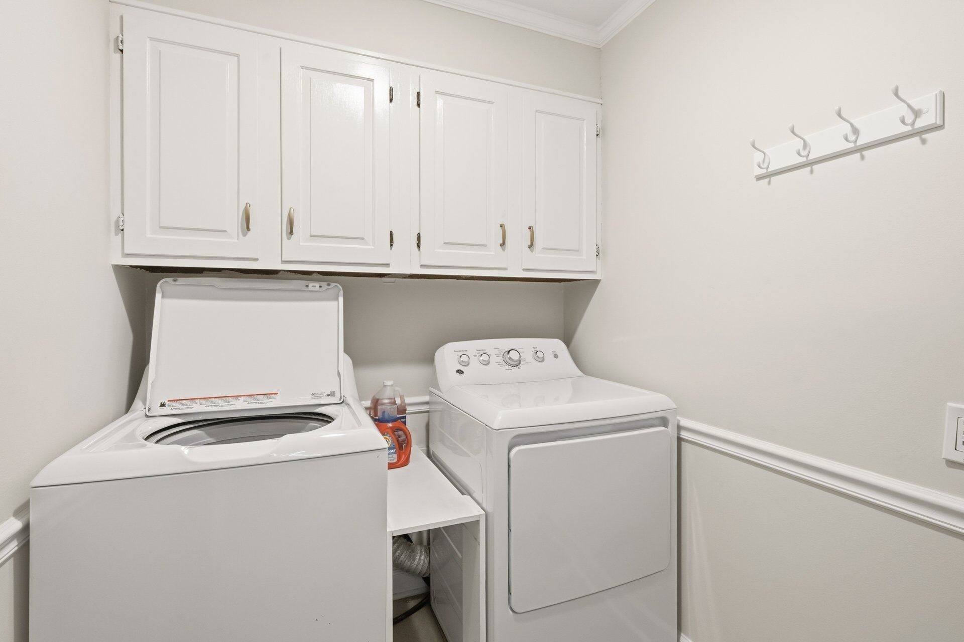 A laundry room with a front-loading washer and dryer, white cabinets above, and a coat rack on the wall.