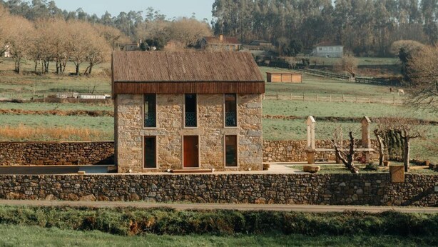 A stone building with a thatched roof, surrounded by a stone wall, set in a rural landscape with fields and trees.