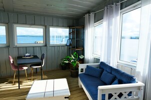 A room with a white sofa, a small table, and two chairs. There are windows with a view of the sea.