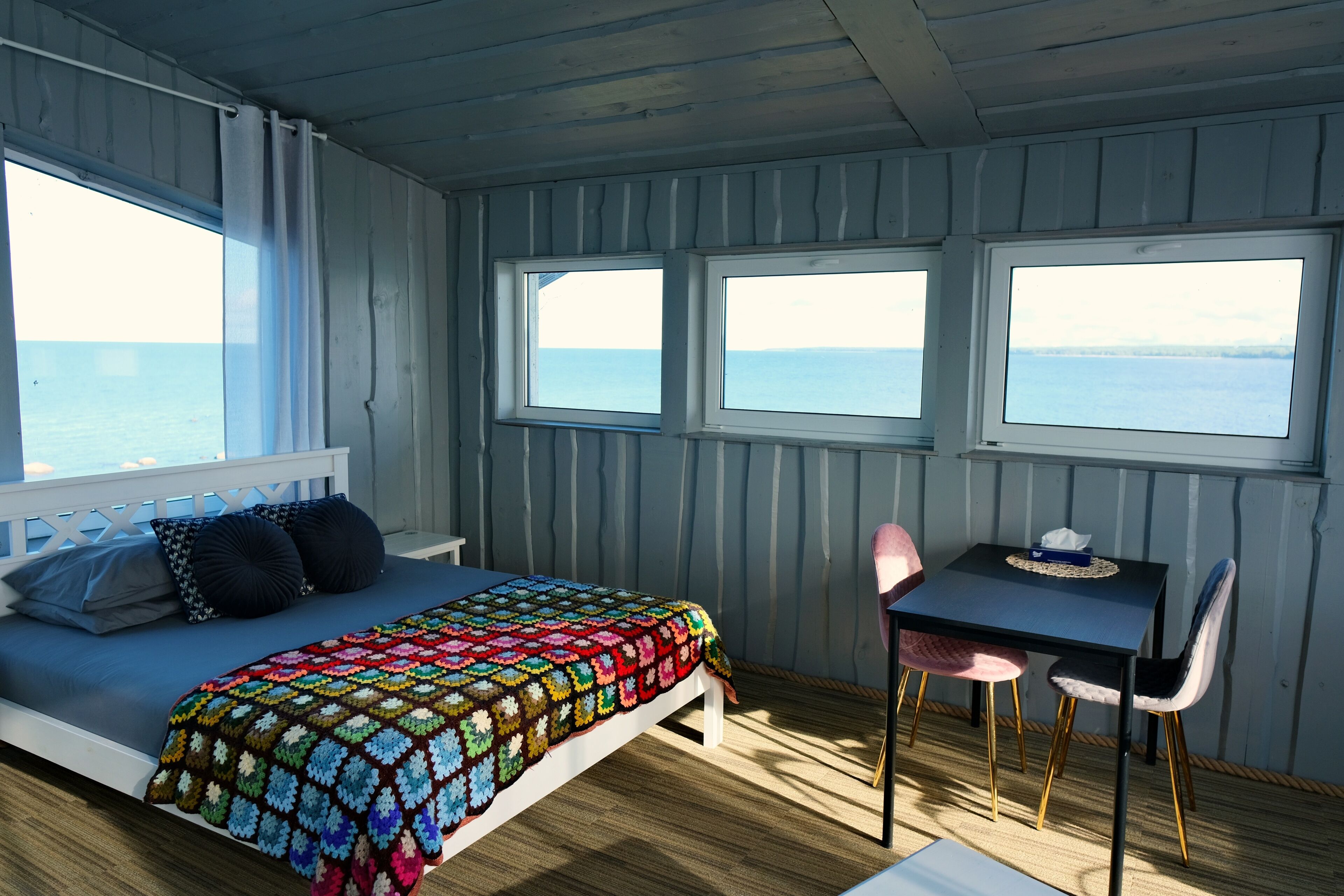 Quarto com cama, mesa e duas janelas com vista para o mar.