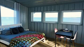 Quarto com cama, mesa e duas janelas com vista para o mar.