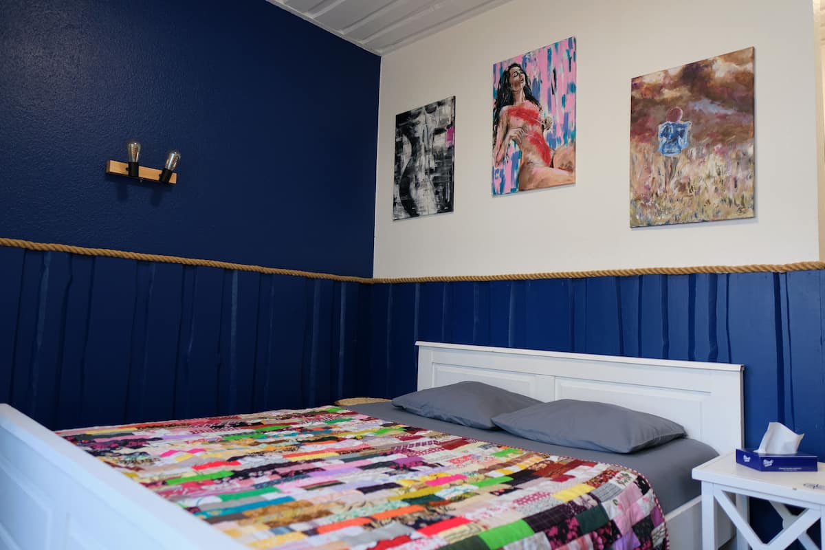 Ein Schlafzimmer mit blauer Wand, einem weißen Bett, einer farbenfrohen Decke und drei Bildern.