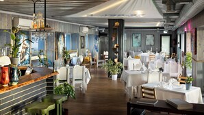 Un restaurant avec un plancher en bois, des tables blanches et des chaises vertes. On y trouve des plantes et un coin bar avec des bouteilles et des verres.