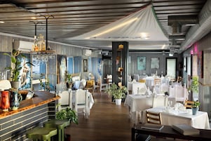 A restaurant with wooden floors, white tables, and green chairs. There are plants and a bar area with bottles and glasses.