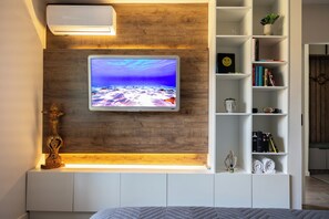 Un dormitorio moderno con un televisor montado, una pared de acento de madera, un estante blanco con libros y objetos decorativos, y una cama con colcha estampada.