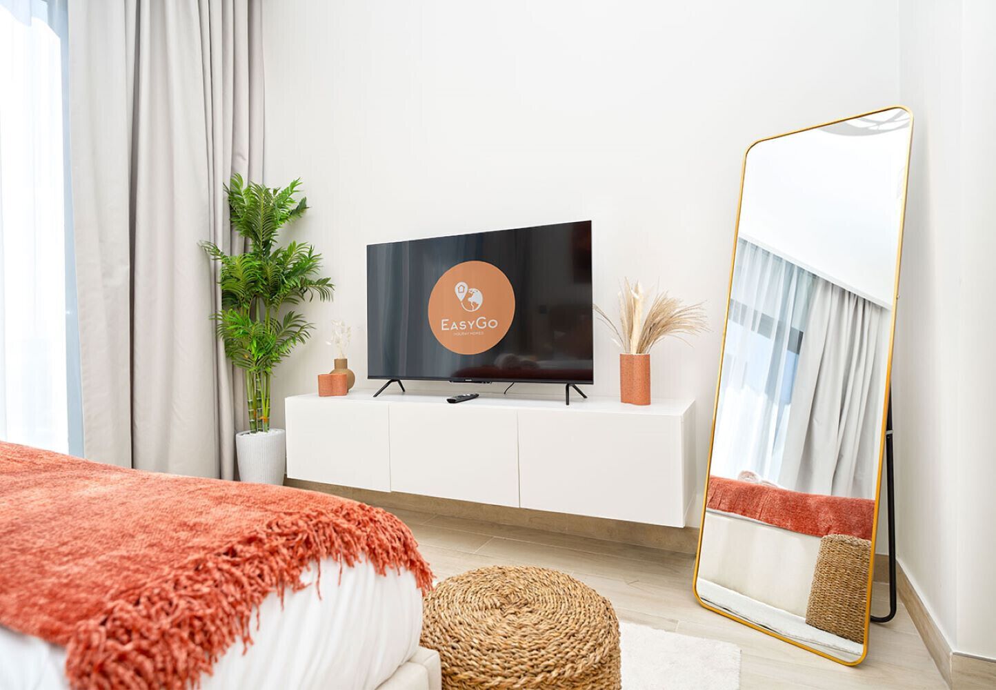 A modern bedroom with a bed, a television on a white cabinet, a large mirror, and a potted plant.