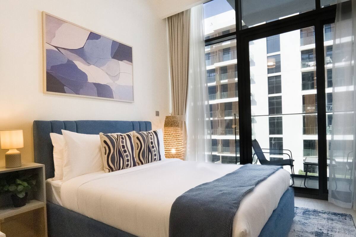 A hotel room with a large bed, a nightstand, a lamp, a plant, a window with a view of a building, and a chair by the window.