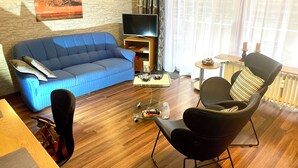 Una sala de estar con un sofá azul, un sillón negro, una mesa de centro de cristal, un televisor y un radiador.