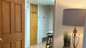 Um corredor com porta de madeira, chão de ladrilho de pedra, uma pequena mesa com luminária e planta, e um banquinho de madeira.
