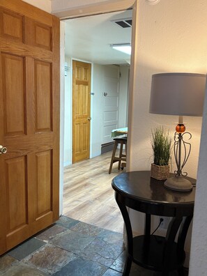 A hallway with a wooden door, a stone-tiled floor, a small table with a lamp and a plant, and a wooden stool.