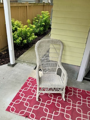 A wicker chair on a red patterned rug outside a house with a wooden fence and green bushes in the background.